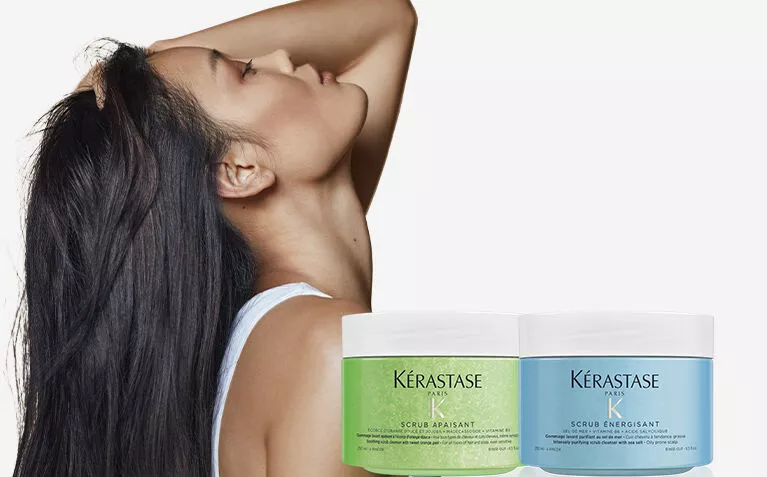 A woman with long, dark hair poses gracefully beside two Kerastase hair scrubs, highlighting hair care and beauty.