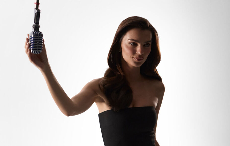 A woman in a black strapless top holds a vintage-style microphone against a plain white background.
