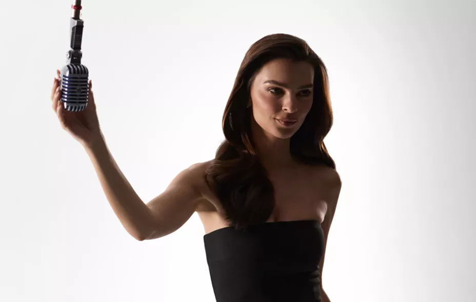 A woman in a black strapless top holds a vintage-style microphone against a plain white background.