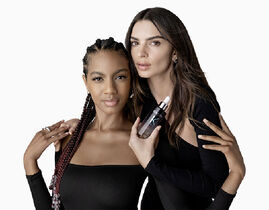 Two women with dark hair pose closely together, one holding a cosmetic bottle. They both wear black outfits and have confident expressions.