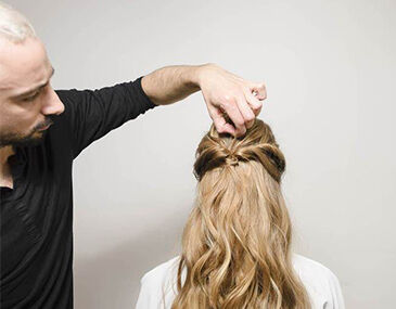 A hairstylist is creating a hairstyle on a person's long, wavy hair from behind, focusing on a section near the crown of the head.