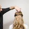 A hairstylist is creating a hairstyle on a person's long, wavy hair from behind, focusing on a section near the crown of the head.