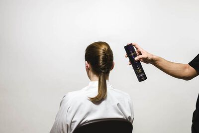 A person with their back to the camera is sitting in a chair while another person is holding a can of hair spray above their head, preparing to style their hair.