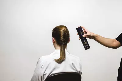 A person with their back to the camera is sitting in a chair while another person is holding a can of hair spray above their head, preparing to style their hair.