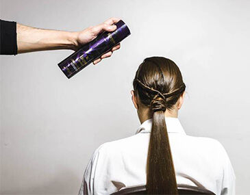 A hand holding a spray can is about to apply hair products to the back of a woman’s long, sleek hairstyle. She is seated and facing away from the camera.