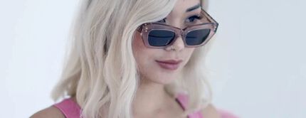 A woman with long, wavy blonde hair wears stylish sunglasses and a pink outfit, exuding a trendy vibe.