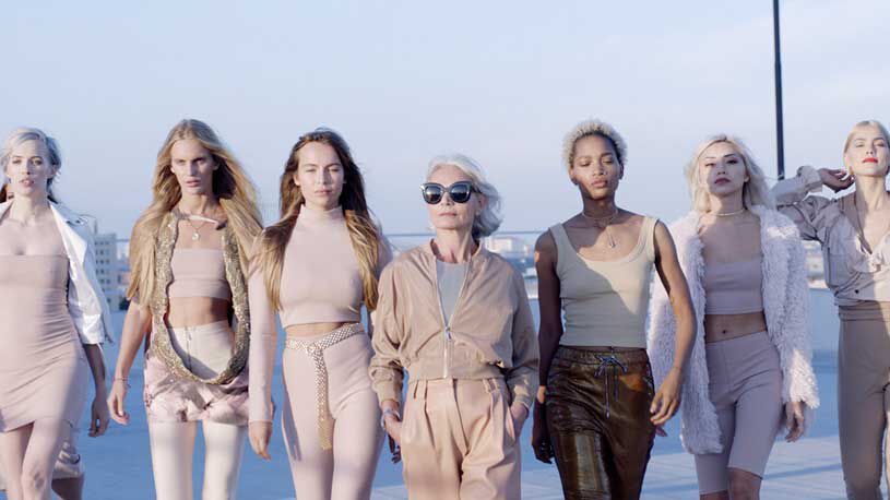 A diverse group of stylish women, including an older model, confidently walk together on a rooftop against a clear sky.
