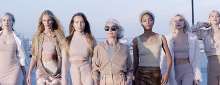 A diverse group of stylish women, including an older model, confidently walk together on a rooftop against a clear sky.