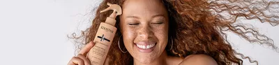 A smiling woman with curly hair and freckles holds a bottle of Kerastase hair product close to her face, showcasing healthy, vibrant hair.