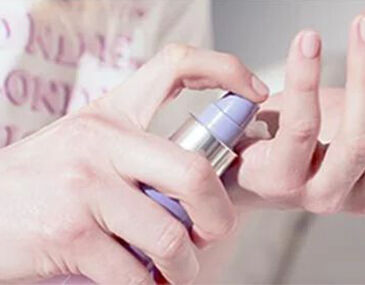 A hand holding a purple pump bottle, dispensing a cream onto a fingertip.
