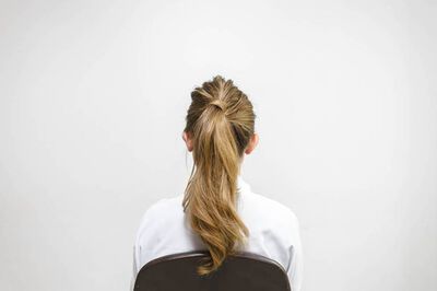 A woman with long, wavy hair is seated, facing away from the camera against a plain white wall. She is wearing a white shirt.