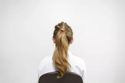 A woman with long, wavy hair is seated, facing away from the camera against a plain white wall. She is wearing a white shirt.