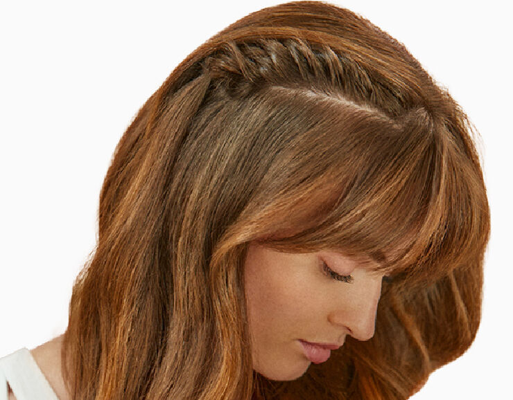 A close-up of a woman with long, wavy brown hair styled with a braided section on one side and soft bangs, looking downward.