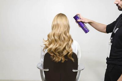 A person is sitting in a salon chair, showing long, wavy blonde hair, while someone off-frame is spraying hair product to style the hair.