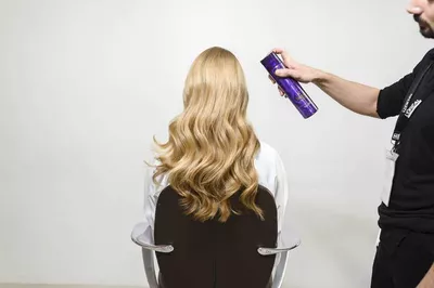 A person is sitting in a salon chair, showing long, wavy blonde hair, while someone off-frame is spraying hair product to style the hair.