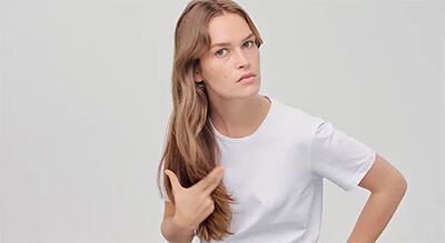 A young woman with long, wavy hair wearing a plain white t-shirt, looking thoughtfully at the camera with a slight expression.