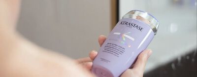 A person holding a bottle of Kerastase purple shampoo with a sleek design and holographic label.
