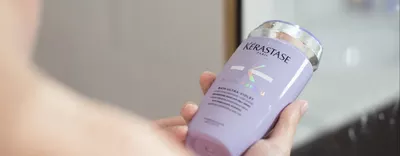 A person holding a bottle of Kerastase purple shampoo with a sleek design and holographic label.