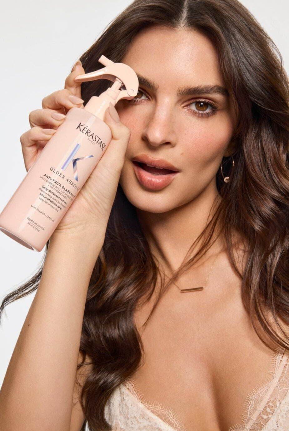 A woman with long, wavy hair holds a bottle of Kerastase hair product close to her face, smiling and looking directly at the camera. She has a natural makeup look and wears a delicate necklace.