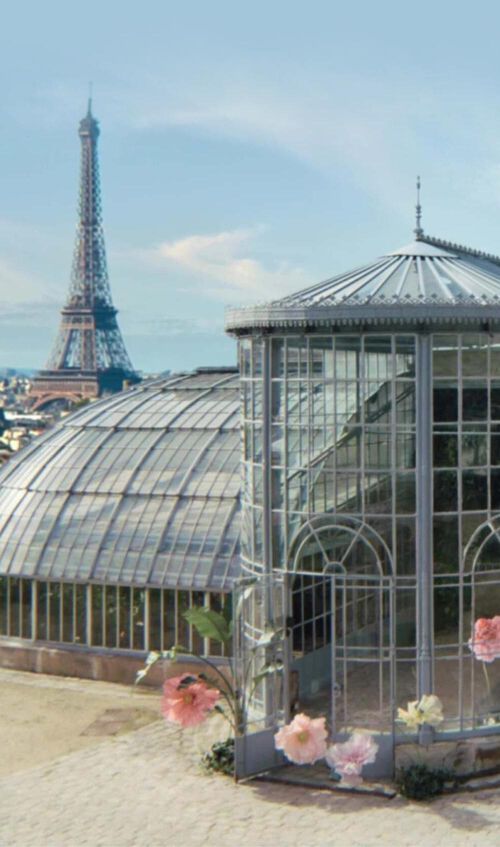 A glass greenhouse with a rounded dome structure, surrounded by a cityscape that includes the Eiffel Tower in the background. Soft pink flowers are partially visible in the foreground.