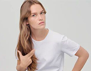 A young woman with long, wavy hair wearing a plain white t-shirt, looking thoughtfully at the camera with a slight expression.