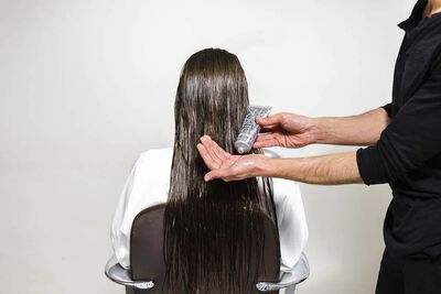 A person with long, wet hair is sitting in a salon chair while a stylist holds a tube of product in one hand and applies it to the hair. The background is plain and white.