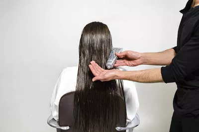 A person with long, wet hair is sitting in a salon chair while a stylist holds a tube of product in one hand and applies it to the hair. The background is plain and white.