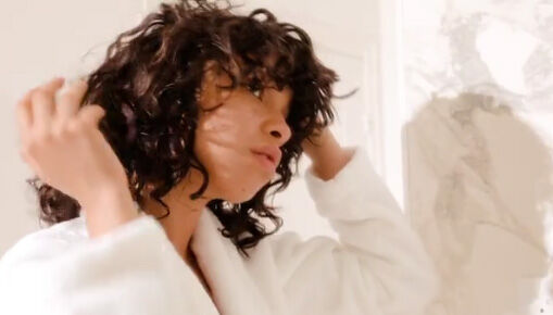A person with curly hair adjusts their hair while wearing a white robe, standing in a bright, elegantly styled room.