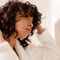 A person with curly hair adjusts their hair while wearing a white robe, standing in a bright, elegantly styled room.