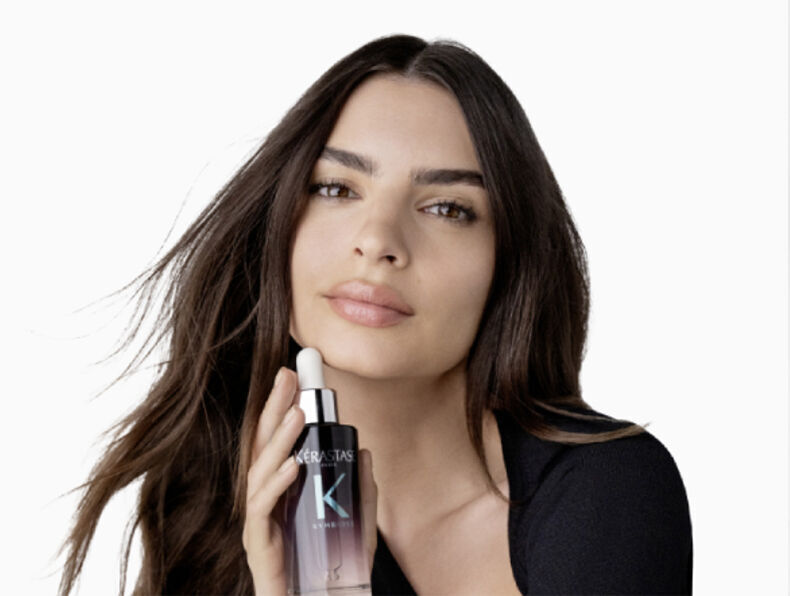 A woman with long, dark hair holds a bottle of Kerastase serum close to her face, promoting a skincare product. She has a soft expression against a plain white background.