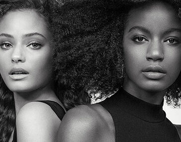 A black and white portrait of two women standing back to back, showcasing different hairstyles—one with wavy hair and the other with voluminous curls. Both have natural makeup, highlighting their features.