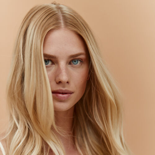 A close-up portrait of a woman with long, wavy blonde hair and striking blue eyes against a warm, peachy background.