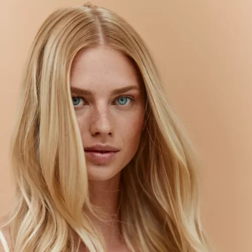 A close-up portrait of a woman with long, wavy blonde hair and striking blue eyes against a warm, peachy background.