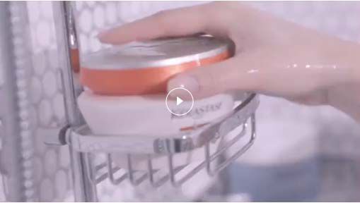 A hand reaching for a round container in a shower caddy with water droplets in the background, suggesting a bath or skincare routine.
