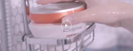A hand reaching for a round container in a shower caddy with water droplets in the background, suggesting a bath or skincare routine.