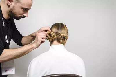 A hairstylist is styling a woman's hair into two elegant bun hairstyles from behind, with the stylist focusing on the details as she sits in a white chair.