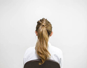 A woman with long, wavy hair is seated, facing away from the camera against a plain white wall. She is wearing a white shirt.