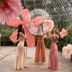 Three women in elegant dresses are lifting a large, transparent sphere in a greenhouse decorated with oversized pink flowers.