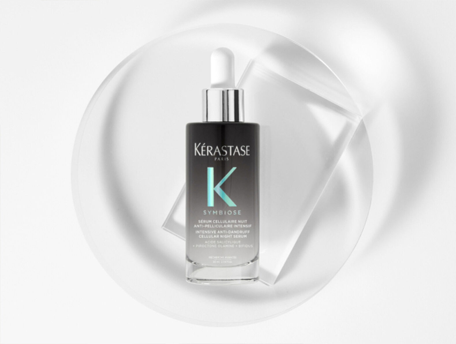 A bottle of Kerastase Symbiose serum displayed on a sleek, white surface with a circular background. The bottle features a dropper and a modern design, indicating a night anti-dandruff treatment.