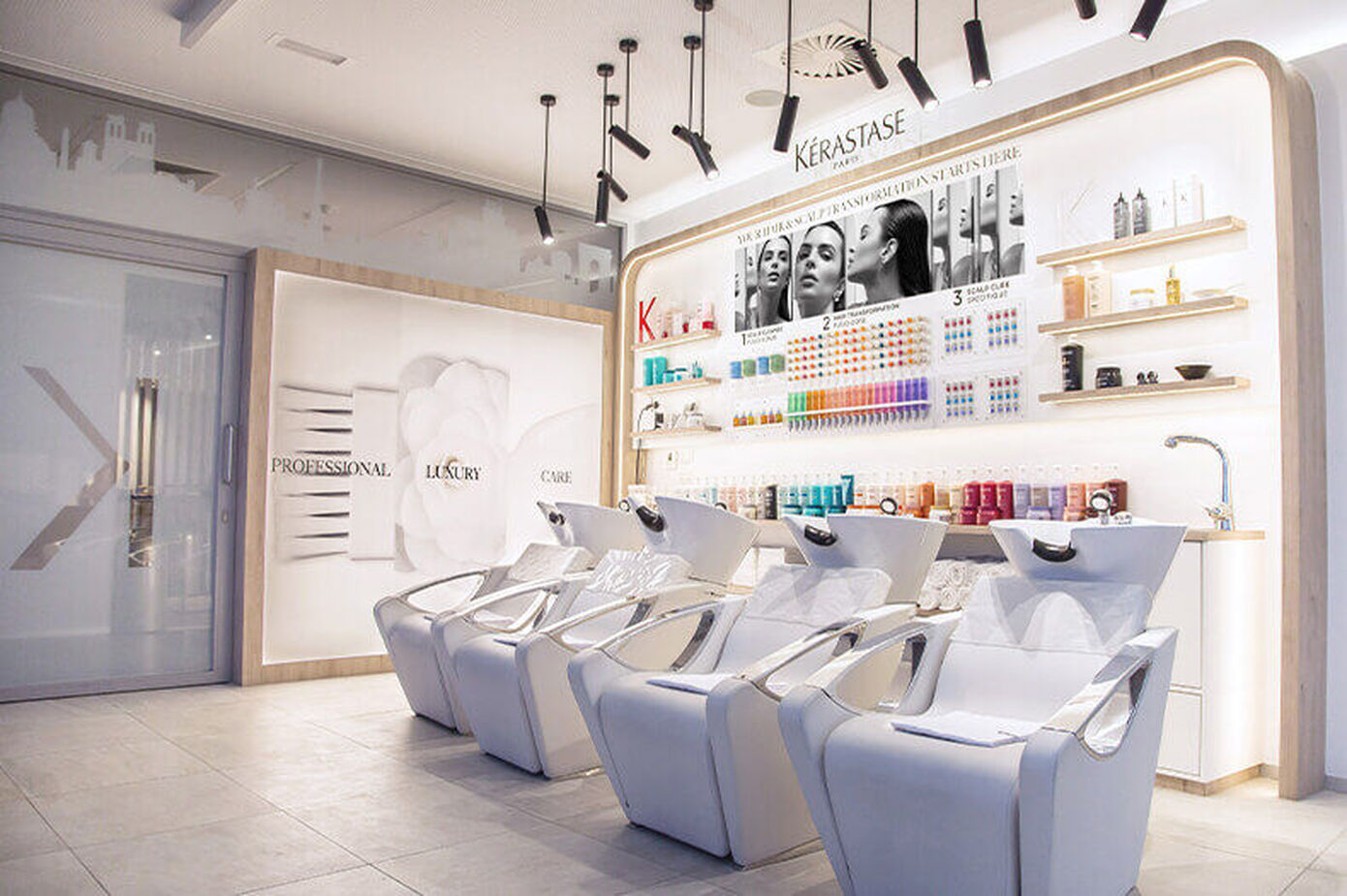 A modern salon interior featuring chic white reclining hair washing chairs and shelves stocked with colorful hair care products against a bright, minimalistic backdrop.