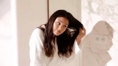 A woman wearing a white robe is brushing her long, dark hair while looking in a mirror. The bathroom has marble accents.