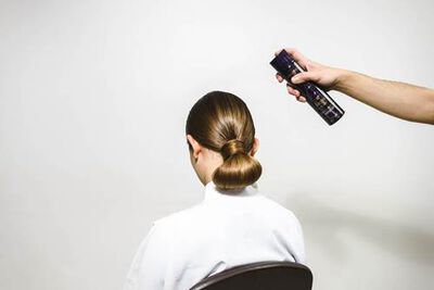 A person sitting in a chair with a sleek bun hairstyle is having hair spray applied from behind.
