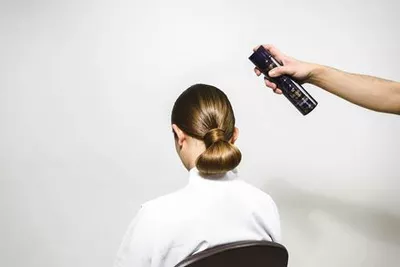 A person sitting in a chair with a sleek bun hairstyle is having hair spray applied from behind.