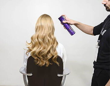 A person is sitting in a salon chair, showing long, wavy blonde hair, while someone off-frame is spraying hair product to style the hair.