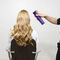 A person is sitting in a salon chair, showing long, wavy blonde hair, while someone off-frame is spraying hair product to style the hair.