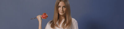A woman with long, straight hair holds a pink rose against a solid blue background, wearing a light-colored top.