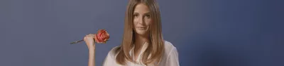 A woman with long, straight hair holds a pink rose against a solid blue background, wearing a light-colored top.