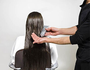 A person with long, wet hair is sitting in a salon chair while a stylist holds a tube of product in one hand and applies it to the hair. The background is plain and white.