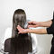 A person with long, wet hair is sitting in a salon chair while a stylist holds a tube of product in one hand and applies it to the hair. The background is plain and white.