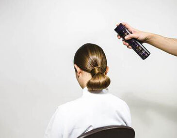 A person sitting in a chair with a sleek bun hairstyle is having hair spray applied from behind.